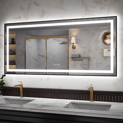 A large rectangular bathroom mirror with a black frame and integrated LED lighting, mounted on a marble-patterned wall above a double sink with gold faucets. The mirror reflects a matching bathroom interior with a shower, sconce, and framed art.
