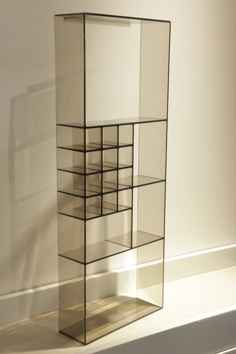 A tall, modern glass shelving unit with a black metal frame and multiple tiers, including a small grid section on the left, set against a warm beige wall.