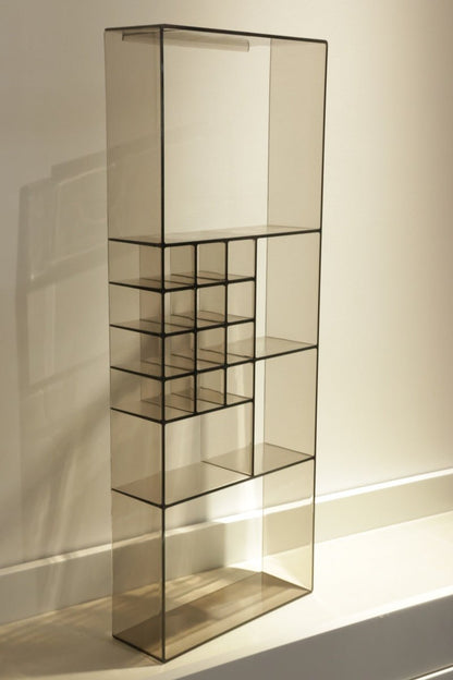 A tall, modern glass shelving unit with a black metal frame and multiple tiers, including a small grid section on the left, set against a warm beige wall.